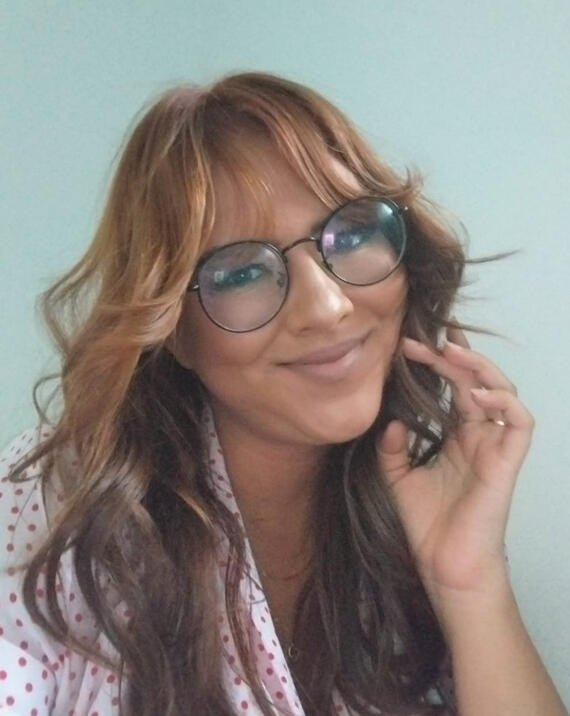 Fotografia de Lolline Huntar’z, uma pessoa não-binária (elu/ela) branca e ruiva com cabelos ondulados, usando um óculos. Seus olhos são castanhos e ela está maquiada, com um batom nude. Ela está com a mão no rosto e o fundo atrás dela é branco azulado.